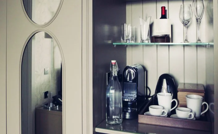 A kitchen cabinet with glassware, a wine bottle, a coffee machine, and cups on a tray.