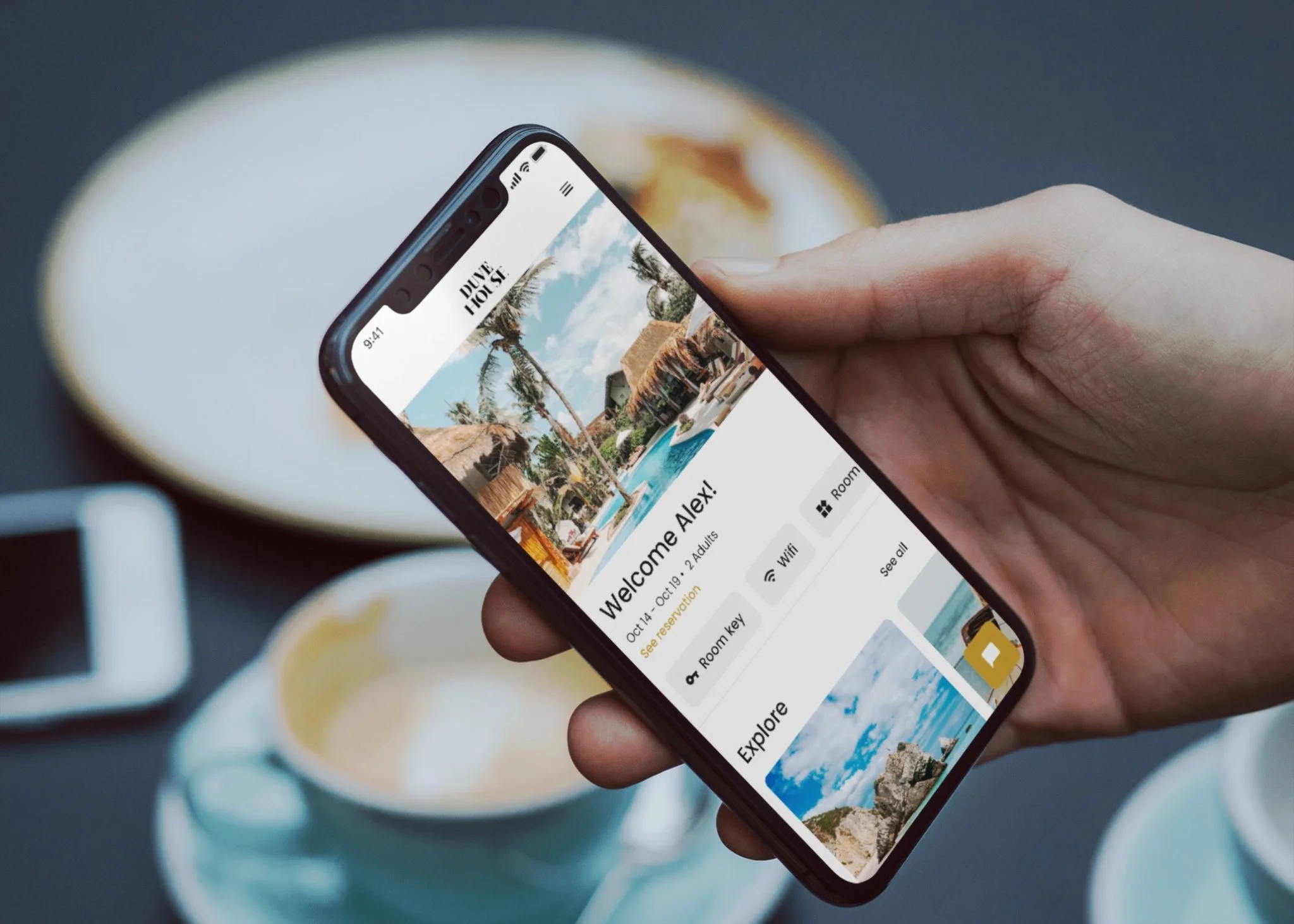 Close-up of a hand holding a smartphone with a travel app displayed, showing a tropical resort with a pool, palm trees, and thatched-roof buildings, with cups of coffee or tea in the background.