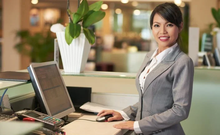 A smiling woman in a gray business suit sitting at a desk with a computer in an office setting.