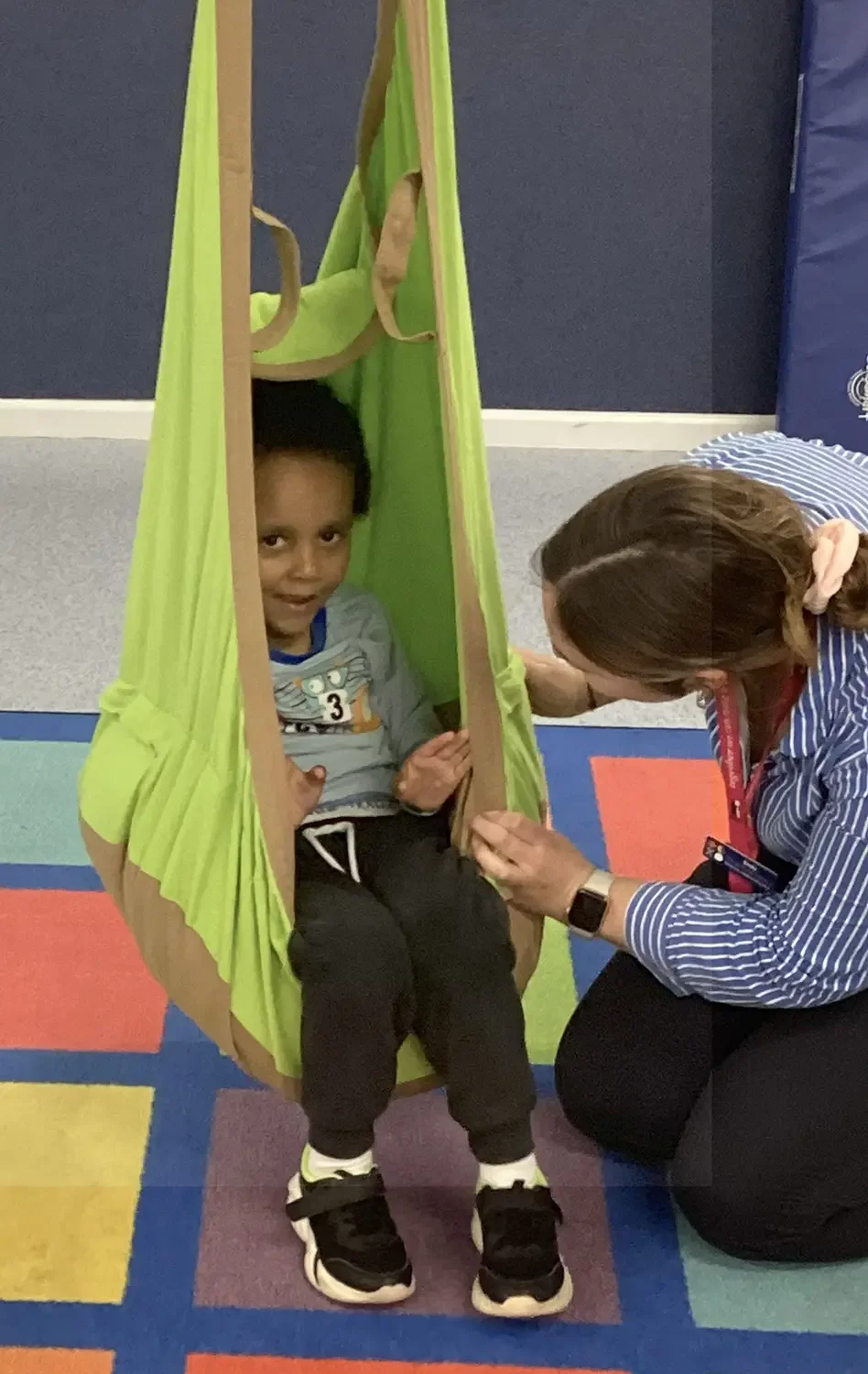 A young boy sitting inside a green fabric hanging chair while a woman kneels beside him, engaging in conversation.