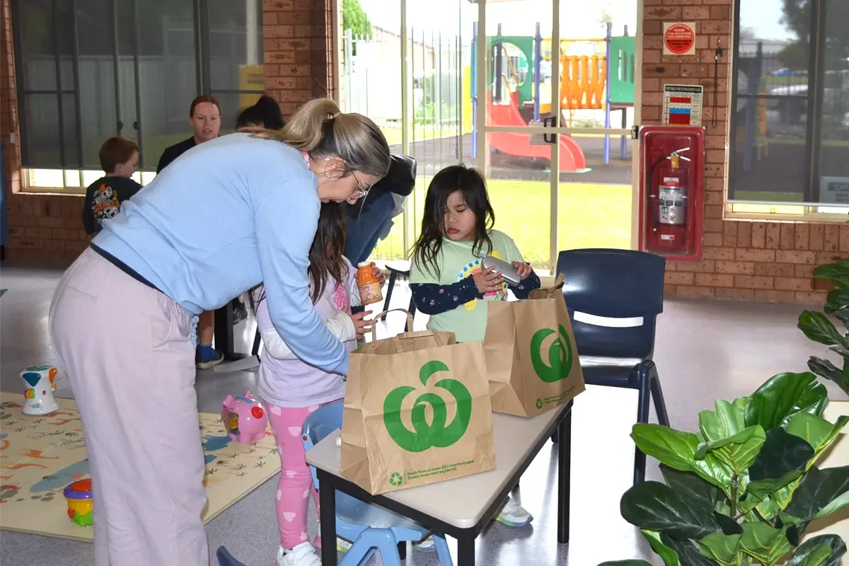 Adults and children at a table entering or collecting items in paper grocery bags in a bright, indoor play area with playground equipment outside.