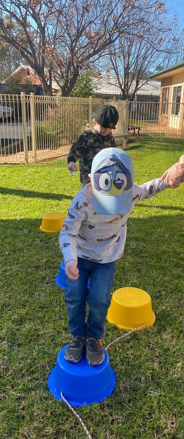 Two young children playing outdoors on a sunny day, with one balancing on a blue plastic bucket and the other in the background. The child in the foreground is wearing a penguin face mask, a long-sleeved shirt with colorful cars, and jeans. The other child wears a camouflage jacket and a black cap.