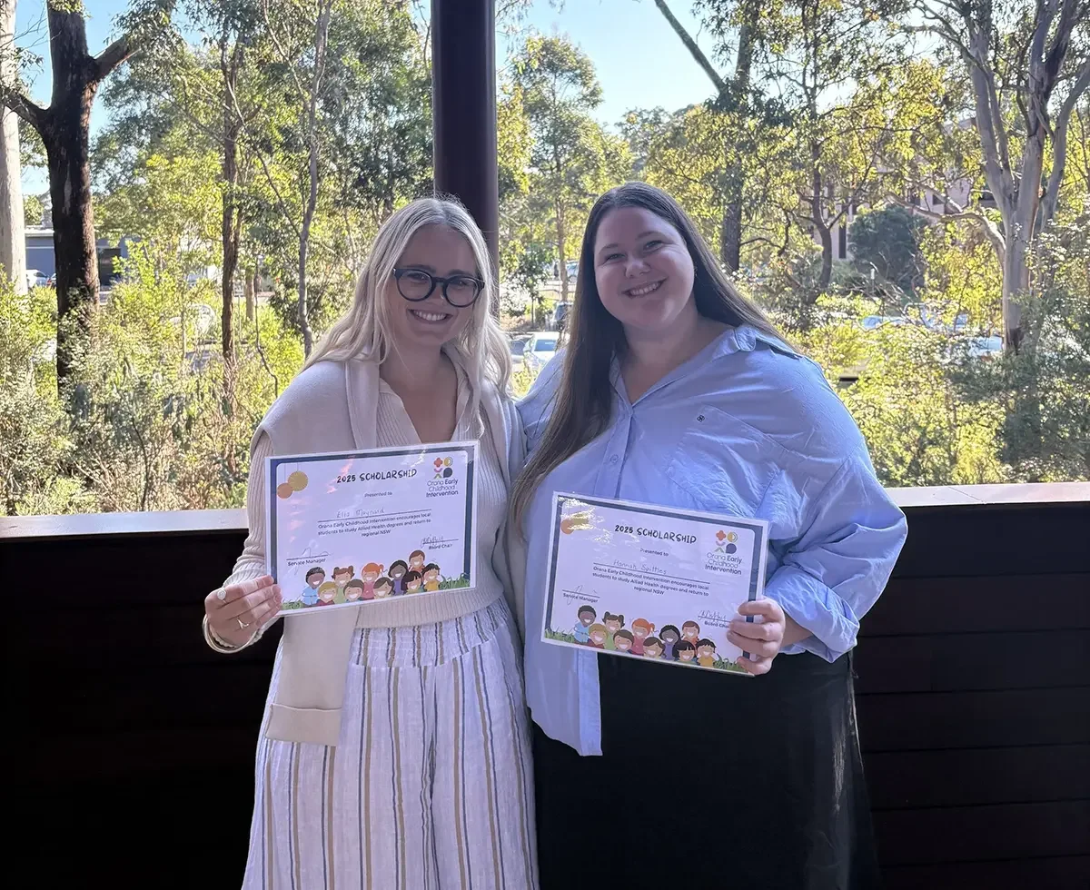 Two women smiling and holding 2023 scholarship certificates, standing outdoors with trees and sunlight in the background.