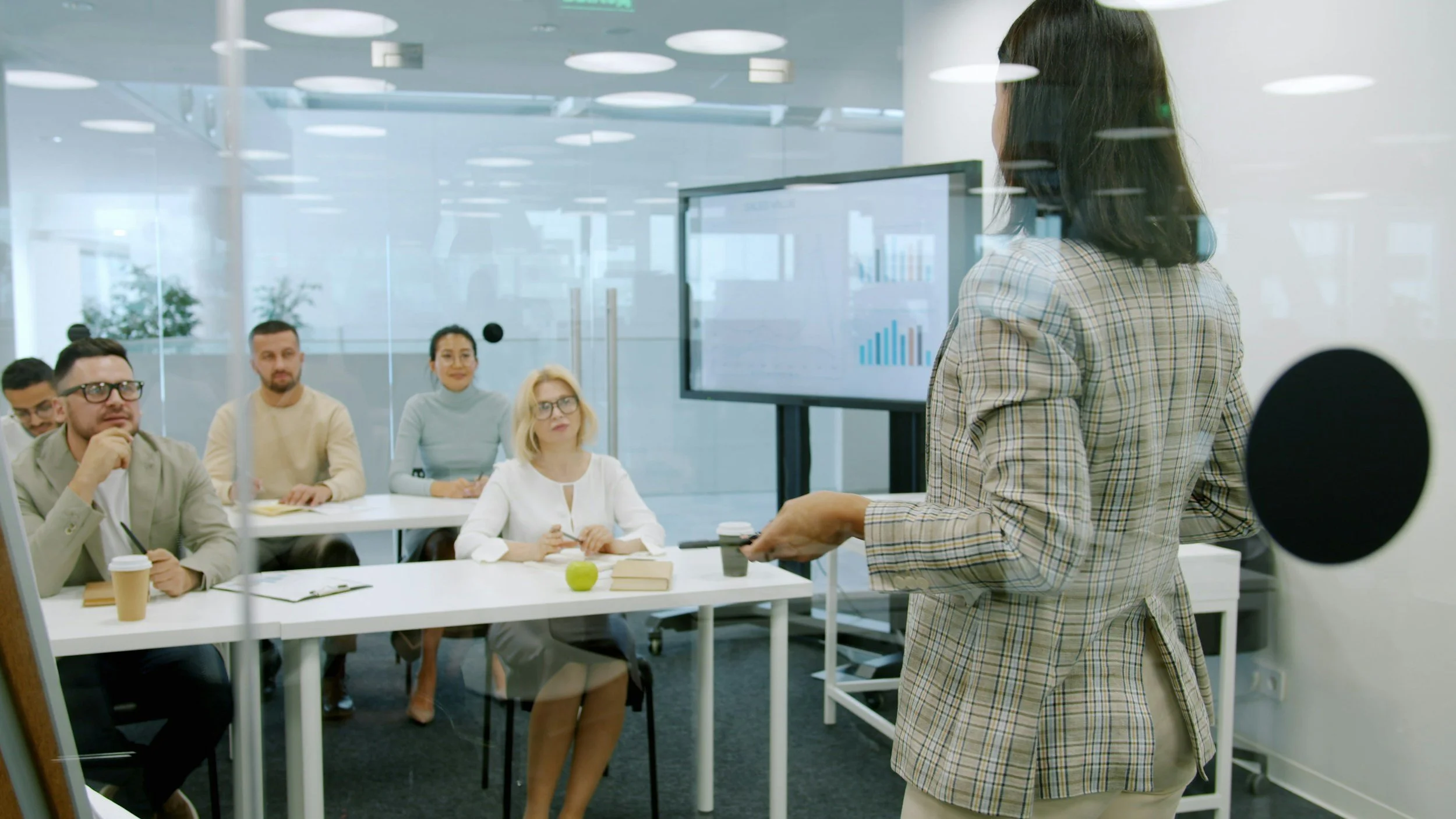 A woman giving a presentation to a group of five people in a conference room with a large screen displaying bar charts, seen through a glass wall.