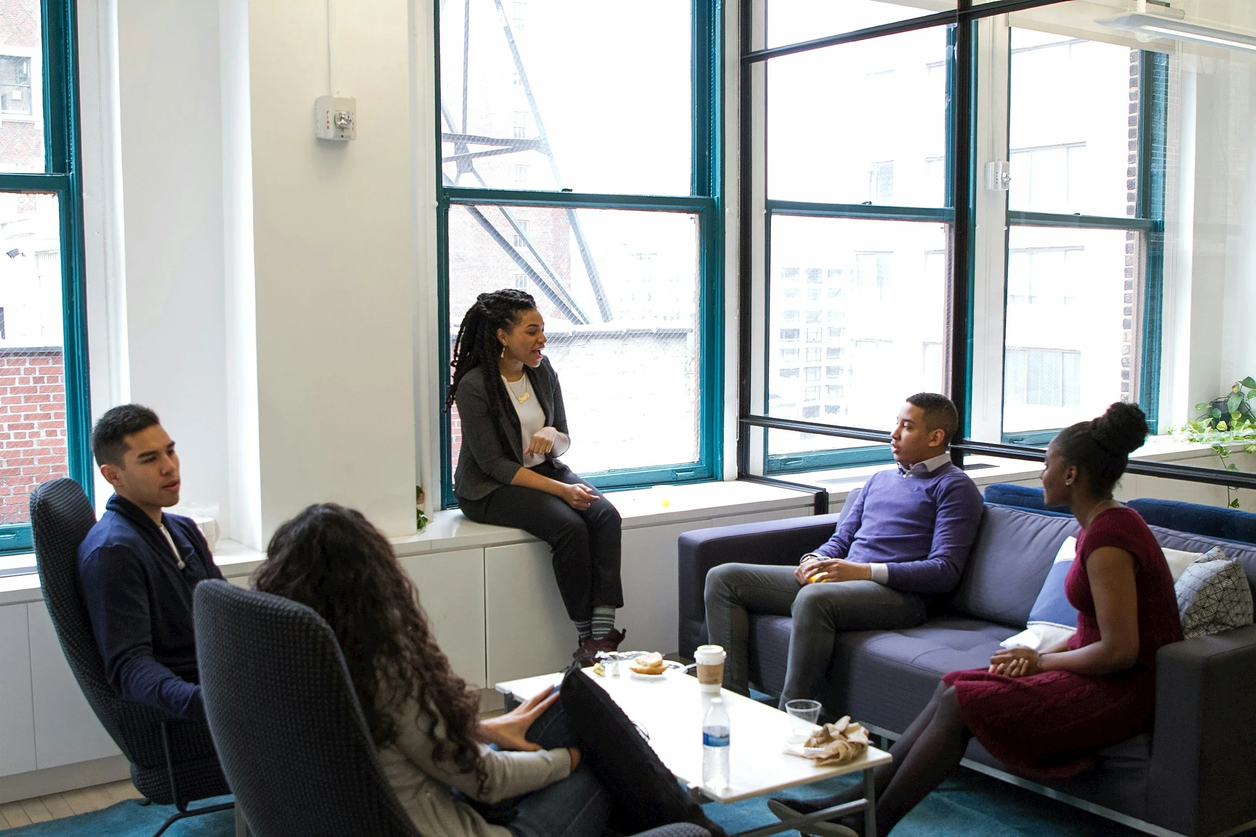 A diverse group of five people having a meeting in a bright office with large windows. One woman is sitting on a windowsill, speaking, while the others are seated around a coffee table listening.