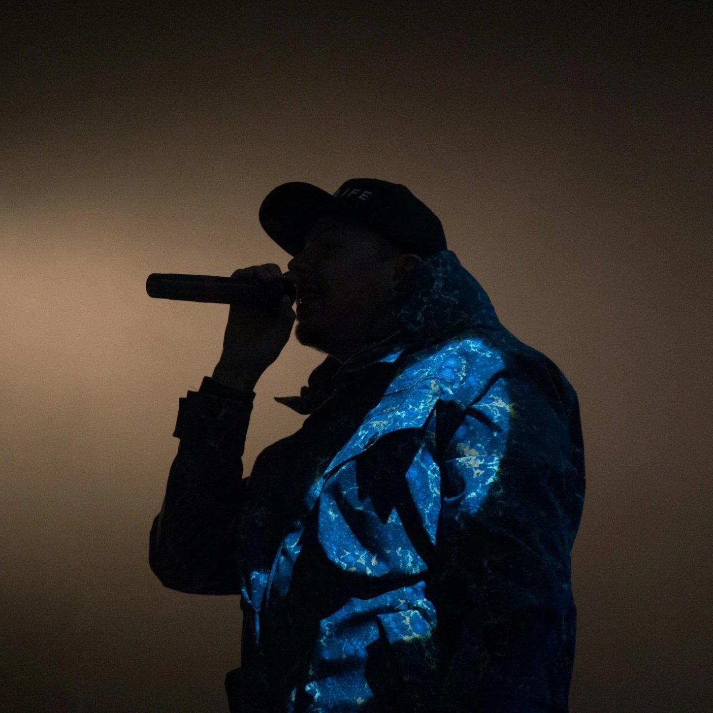 Silhouette of a person holding a microphone, wearing a cap and a jacket with a blue and black pattern, against a dark background.