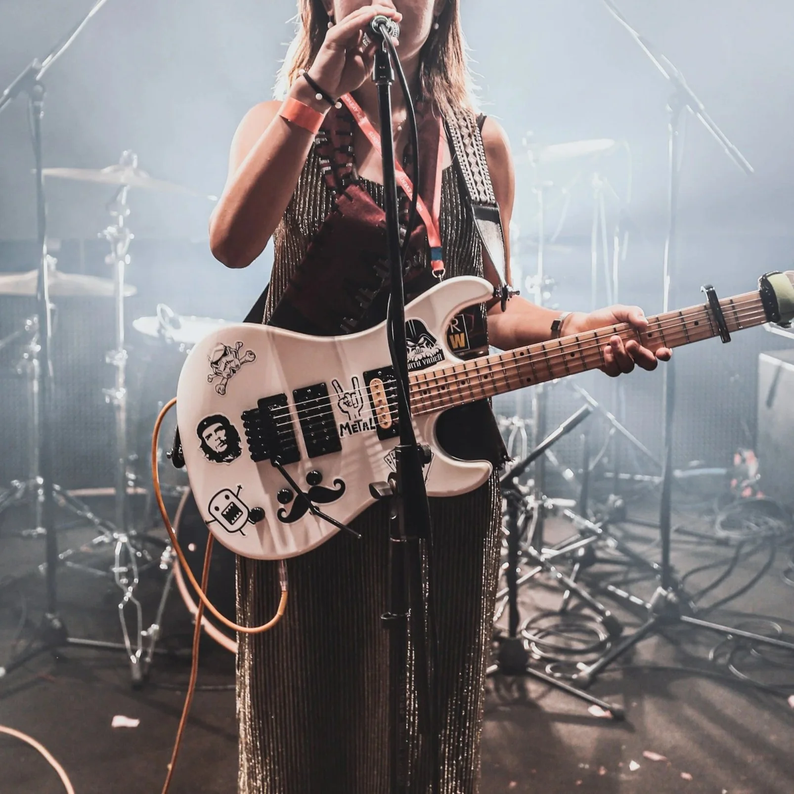 A female musician on stage playing an electric guitar covered in stickers, with a drum set and fog in the background.