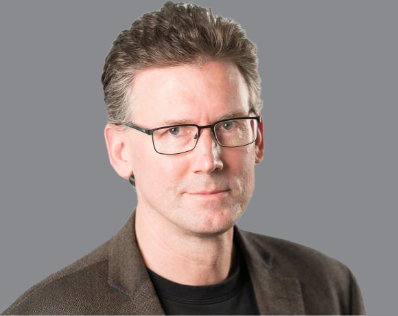 A man with glasses, short brown hair, wearing a dark blazer and black shirt, standing against a gray background.