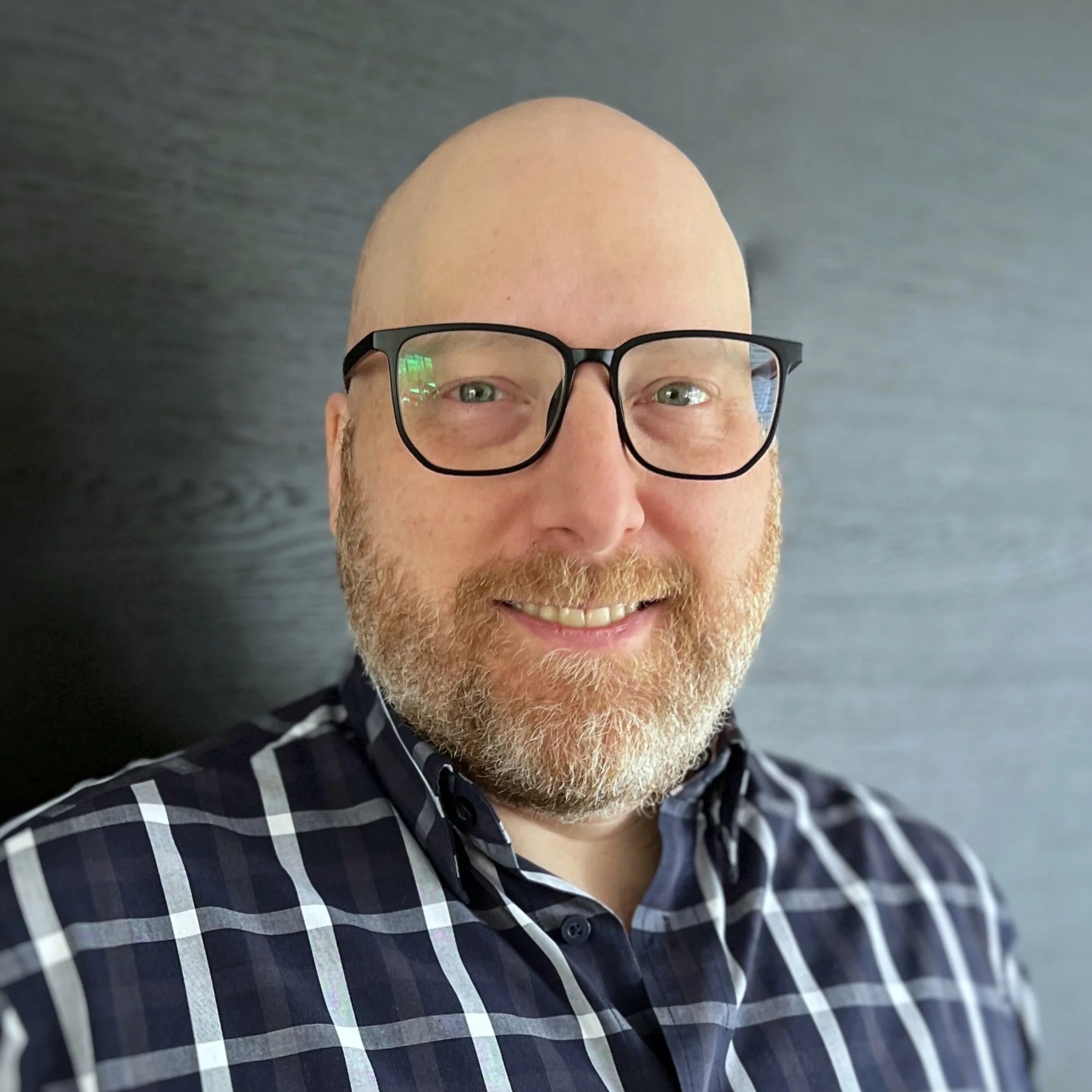A portrait of a man with glasses, a beard, wearing a black and white plaid shirt, smiling against a dark background.