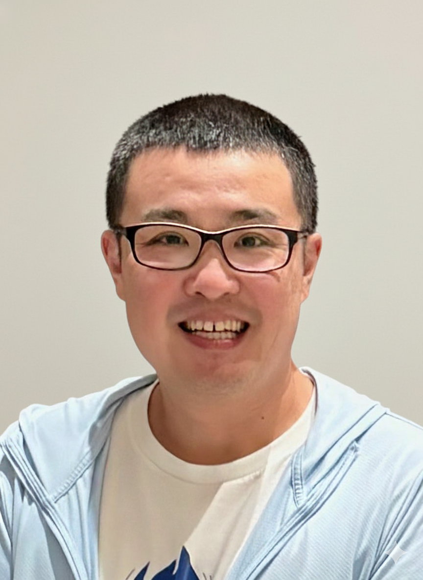 A smiling man with glasses and short dark hair, wearing a white t-shirt and a light blue jacket, standing against a plain background.