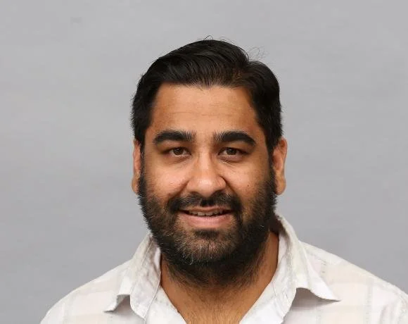 A man with dark hair and a beard smiling at the camera, wearing a white shirt, against a plain gray background.