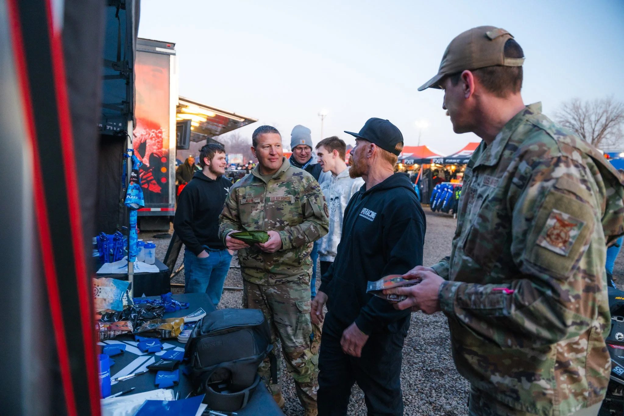 Group of people, including military personnel and civilians, conversing at an outdoor event with tents and merchandise stalls in the background.