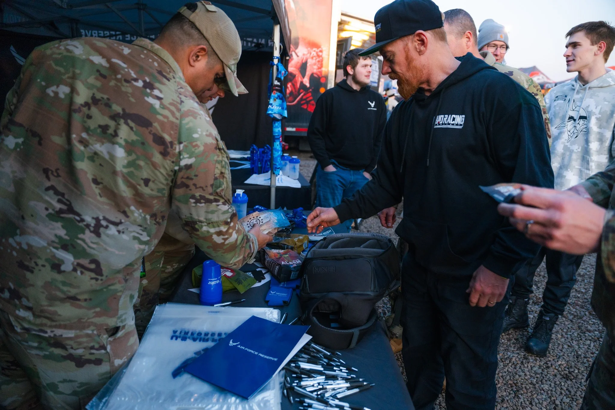 Military personnel at an outdoor event interacting with civilians, with tables displaying promotional items, pens, and blue merchandise, under a canopy during sunset.