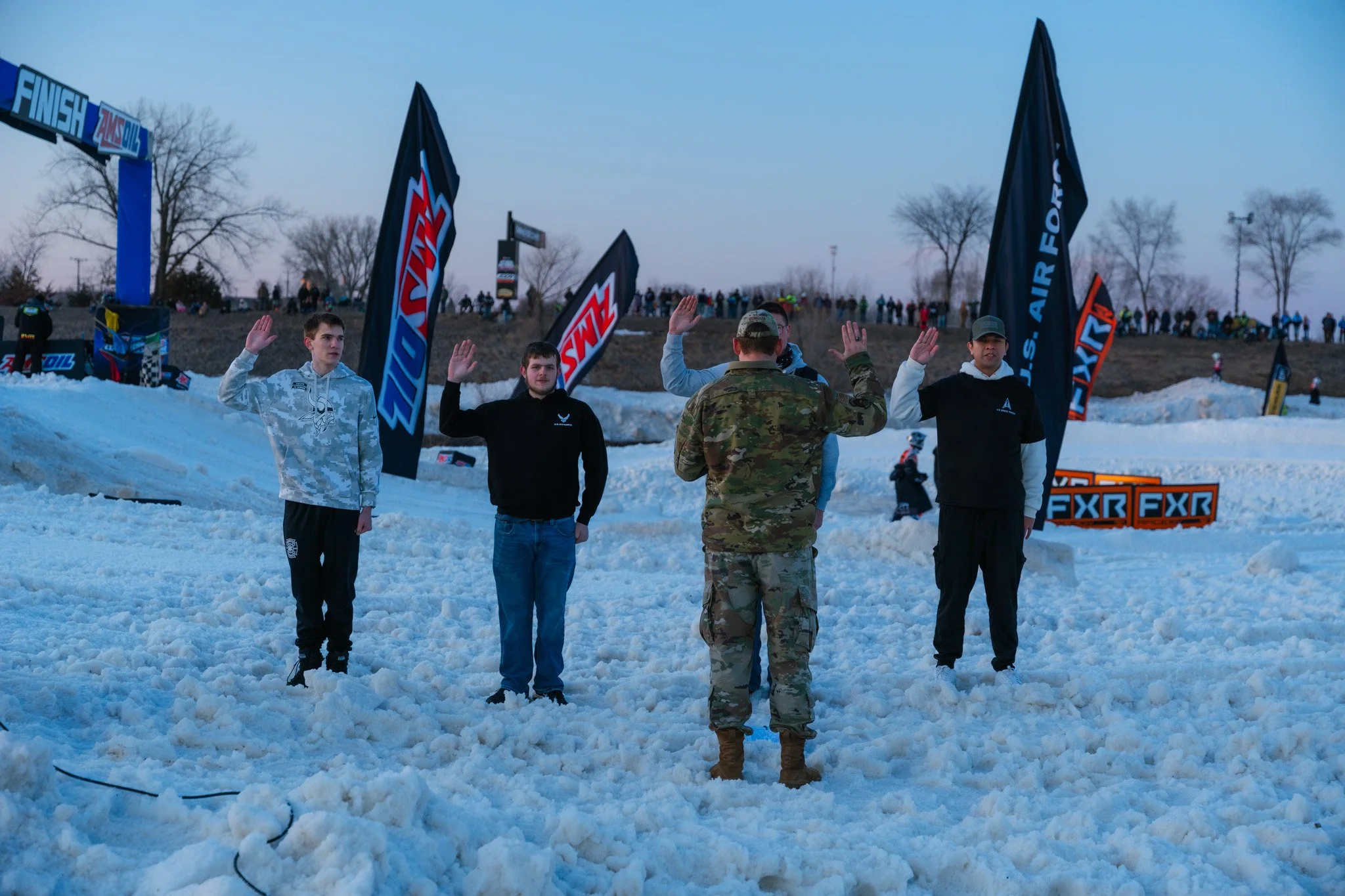 Group of five young men and one military personnel giving a five in the snow at a snow racing event, with race banners and spectators in the background during dusk.