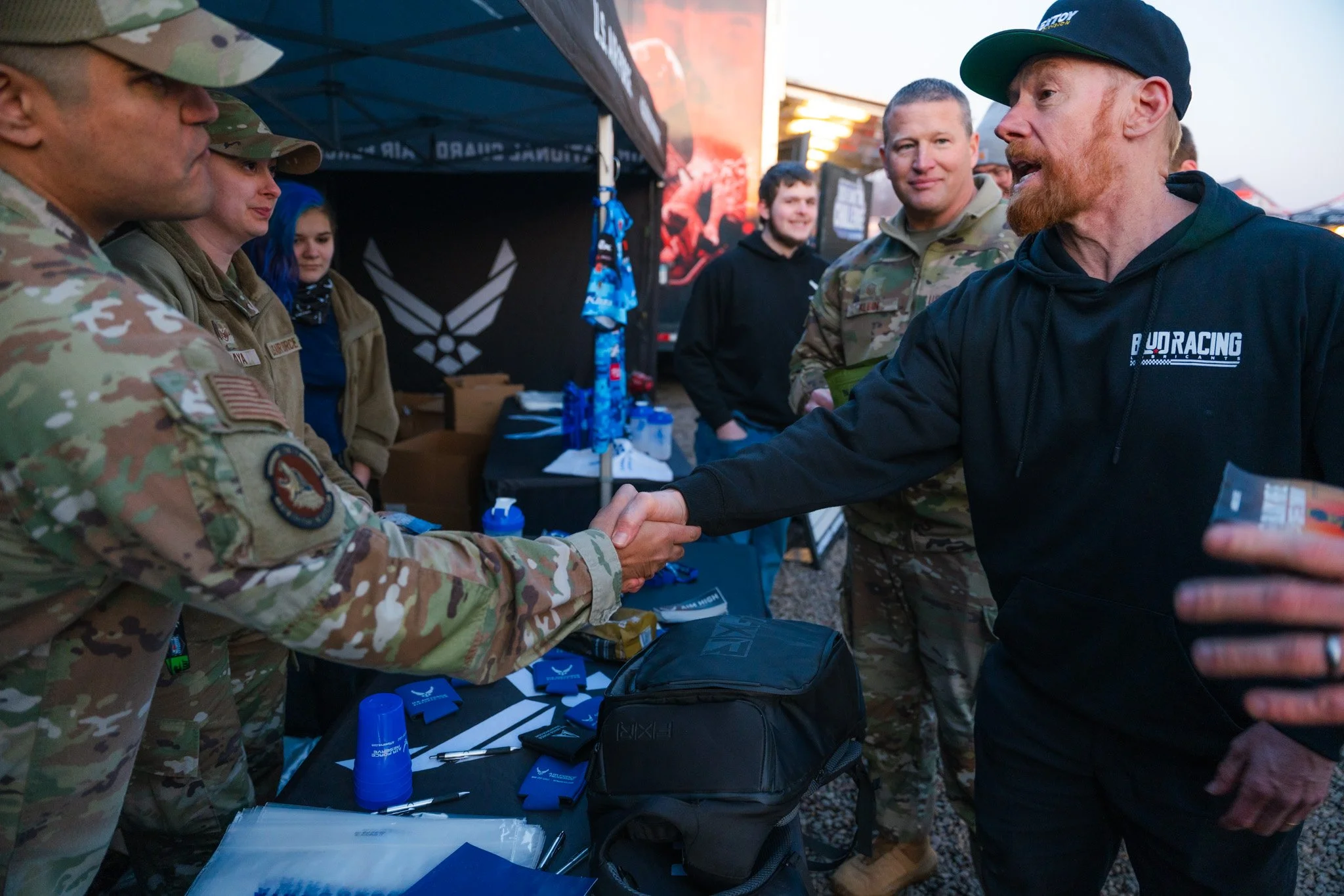 A man with a red beard wearing a black hoodie and cap shaking hands with a soldier in camouflage uniform at an outdoor event. Several other people in camouflage are present, standing by a table with promotional items and a black backpack.