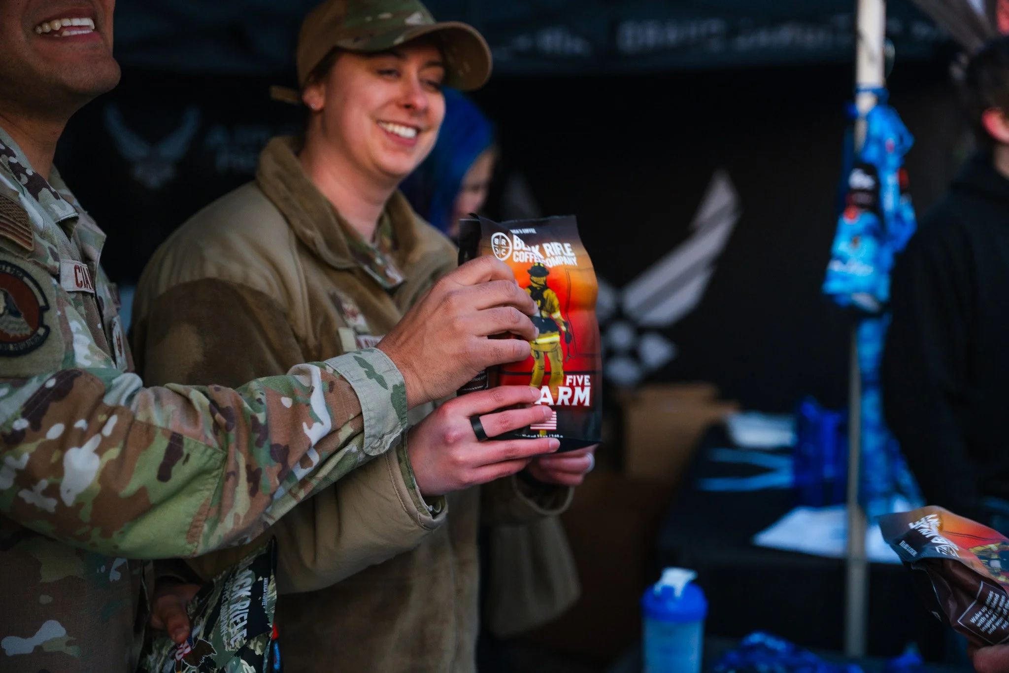 A soldier and a woman in military gear sharing a smile while holding a bag of Five Arm coffee in a casual setting, possibly outdoors or at an event.