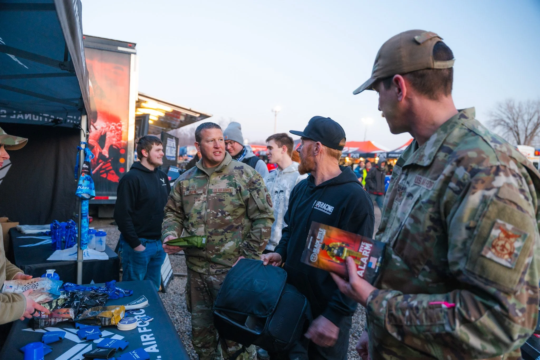 Group of people, including military personnel and civilians, socializing at an outdoor event with tents and trailers in the background during dusk.