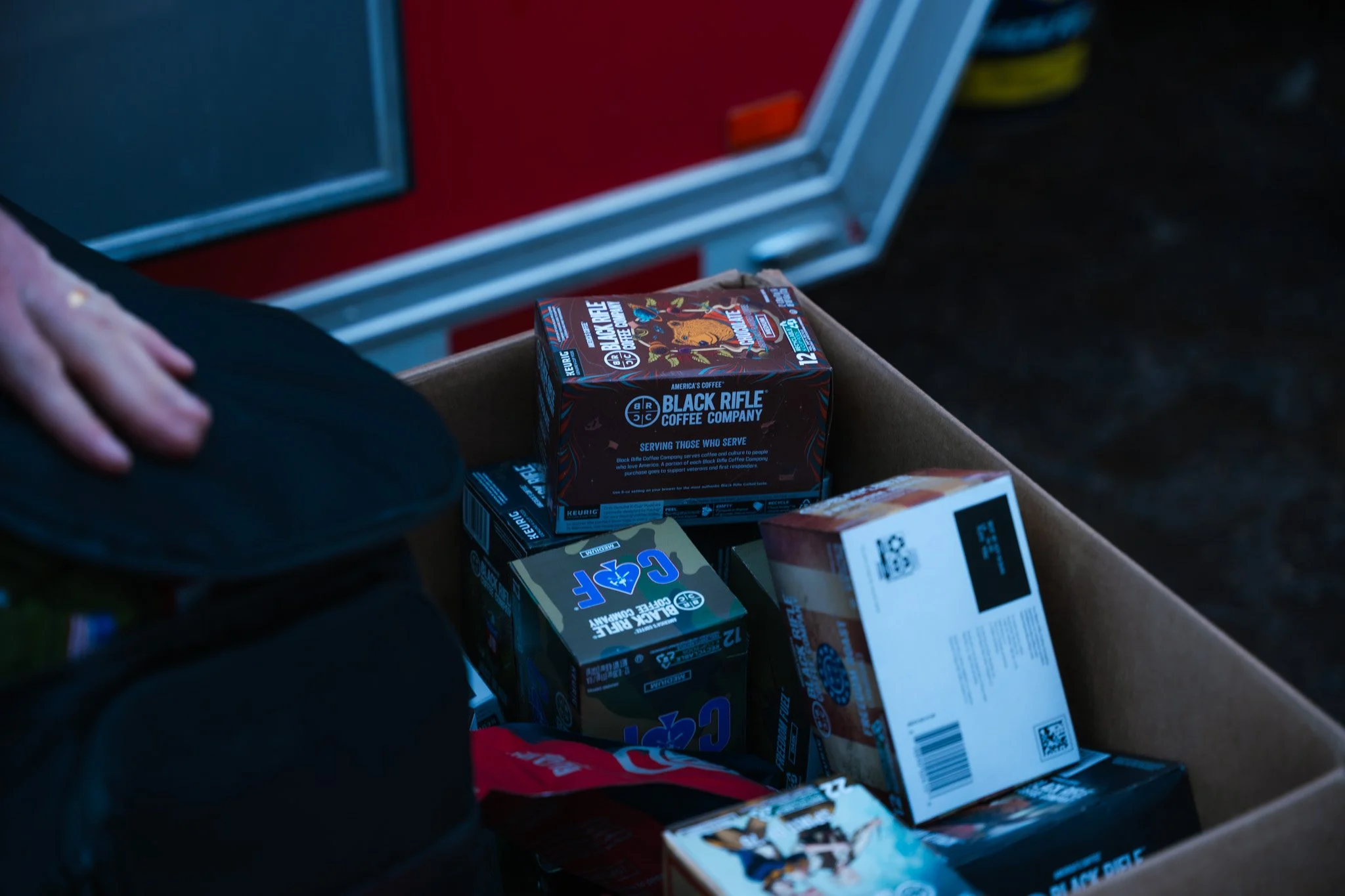 A cardboard box containing various snack and beverage boxes, including a Black Rifle Coffee Company box, with a person's hand resting on a black object nearby.