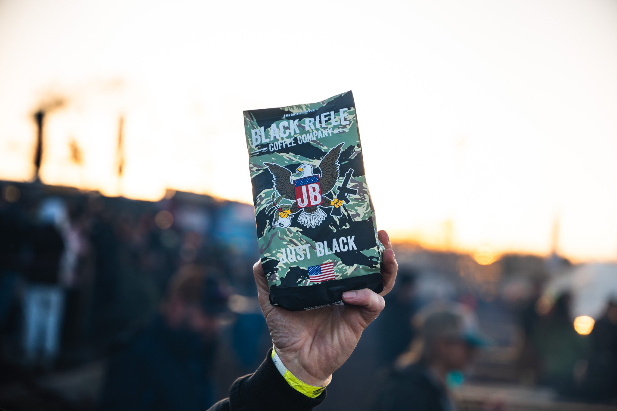 Hand holding a bag of Black Rifle Coffee with a camouflage design and patriotic eagle logo, against a blurred outdoor background at sunset.