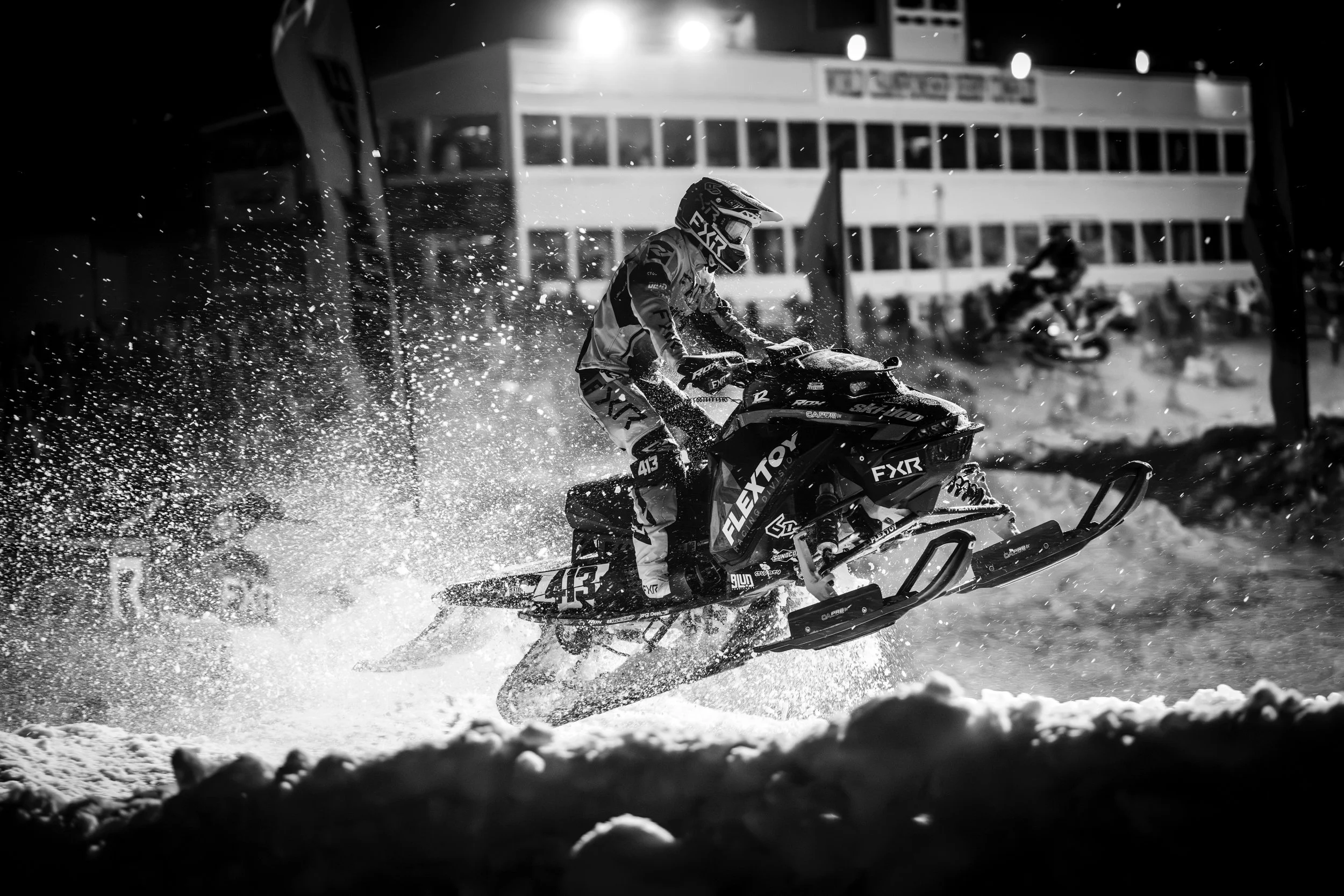 A person riding a snowmobile in mid-air, kicking up snow, with a building and another snowmobile in the background at night.