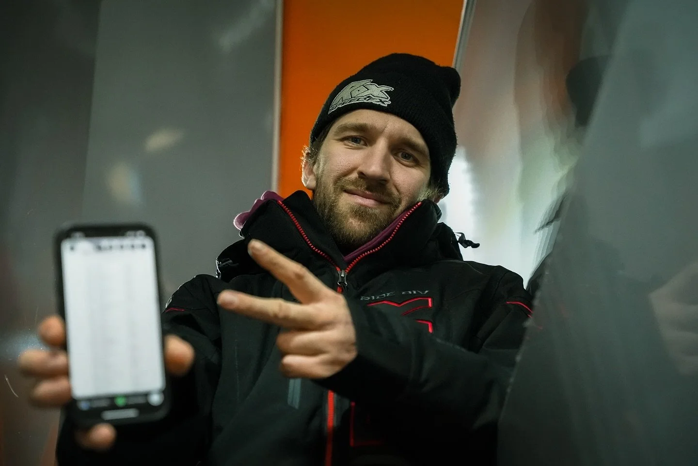 A man wearing a black beanie and black jacket is smiling and making a peace sign with his hand while holding a smartphone in front of him.