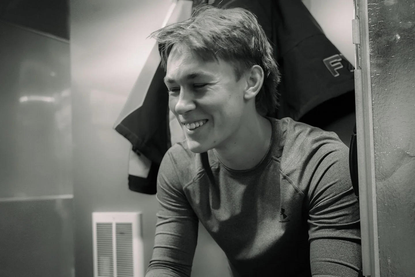 A young man smiling with eyes closed, sitting inside a locker room with a jacket hanging behind him.