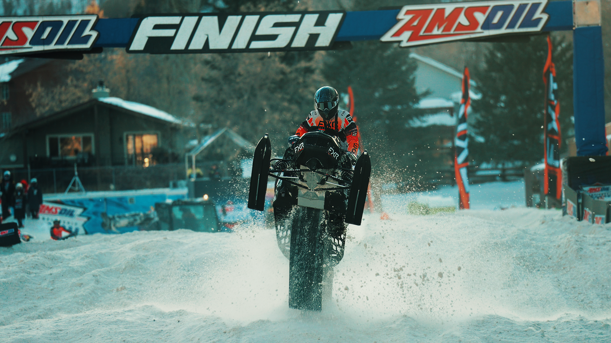 A person riding a snowmobile on snow-covered ground, passing under a finish line with a banner that reads 'FINISH' and 'AMSOIL' logos.