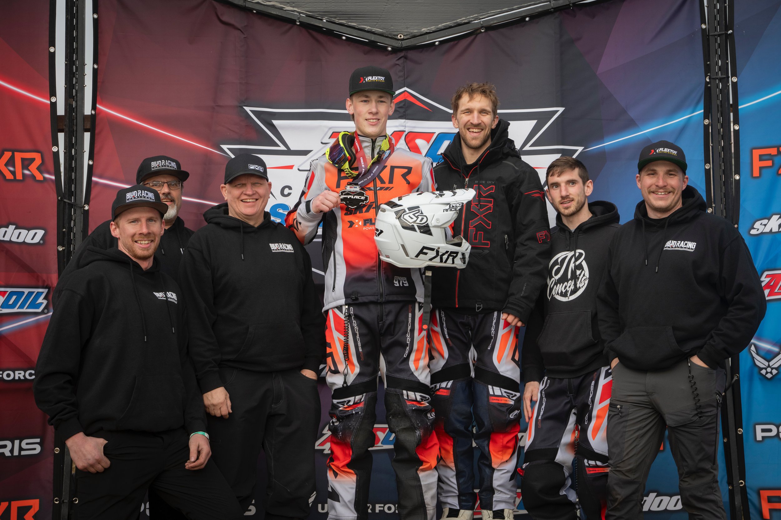 Group of people on a podium celebrating a racing win, with two racers in racing suits holding a helmet and a trophy, surrounded by team members in black hoodies and caps.