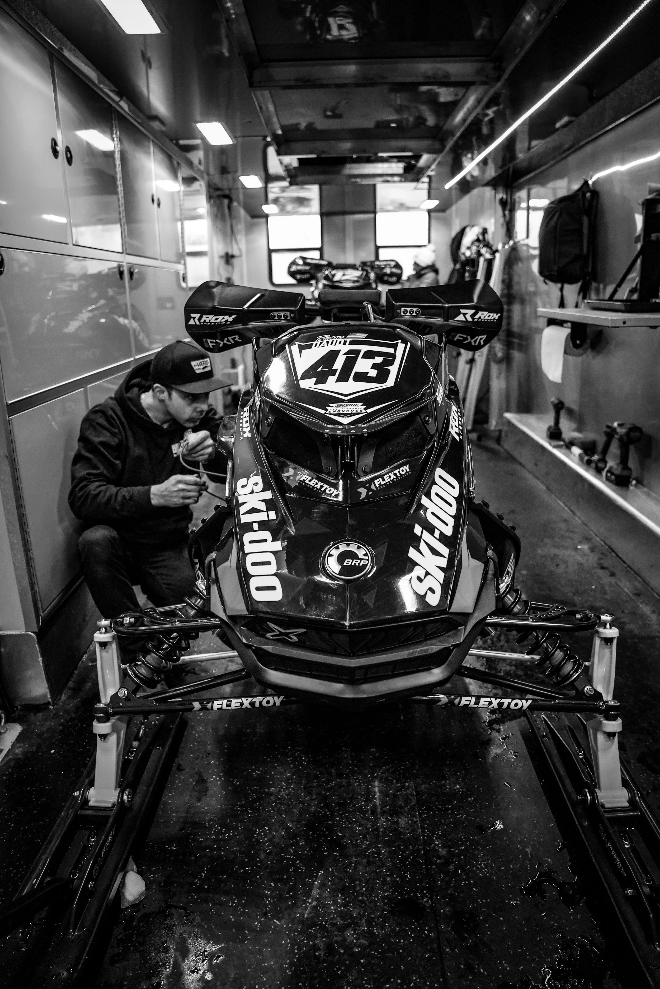 Black and white photo of a racing snowmobile inside a trailer, with a man working on it, surrounded by storage lockers and tools.