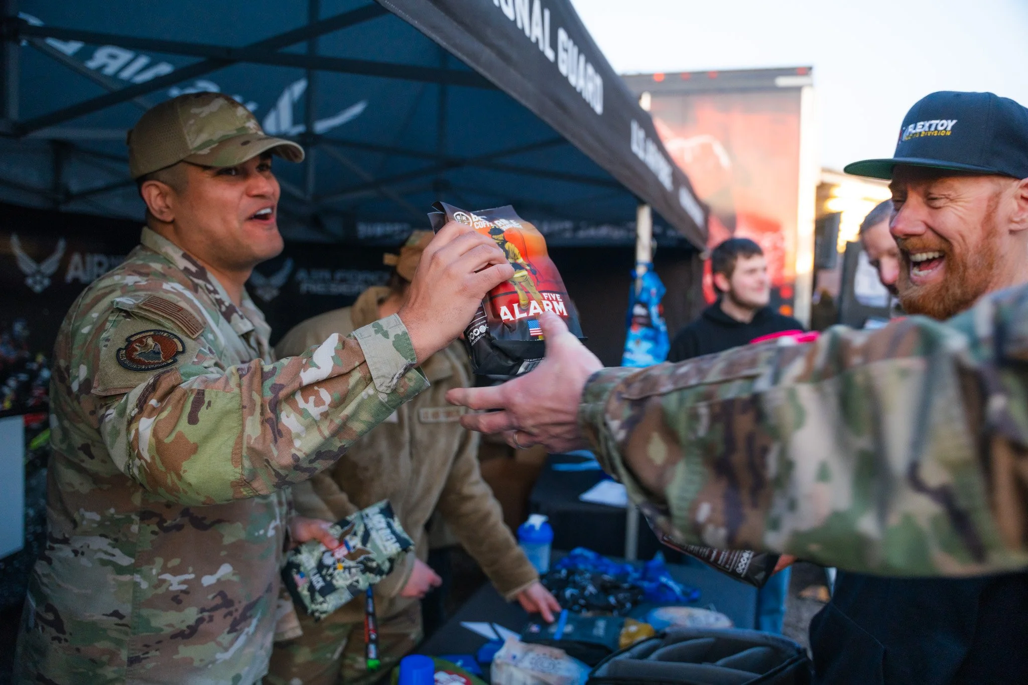 Soldiers in camouflage uniform receiving a bag of snacks and a five alarm fire alarm energy drink at event booth.