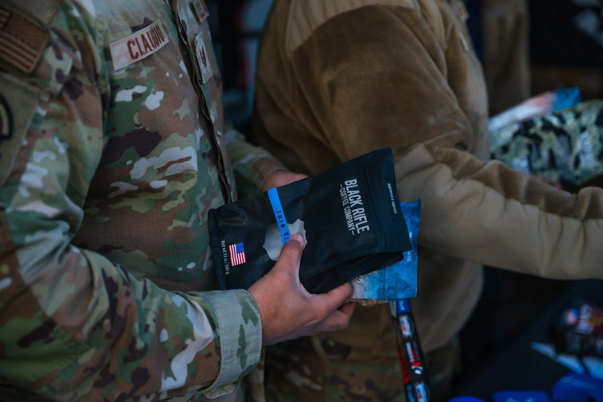 Person in camouflage military uniform holding a bag of Black Rifle Coffee Company thin blue line coffee.