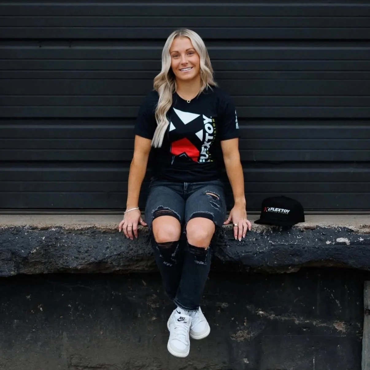 Smiling young woman with long blonde hair, wearing a black T-shirt with a logo, ripped jeans, and white sneakers, sitting on a concrete ledge in front of a black metal door, with a black cap beside her.