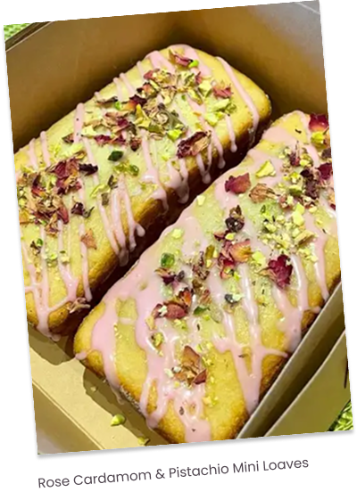 Two slices of cake with pink icing, sprinkled with crushed pistachios and dried rose petals, in a cardboard box.