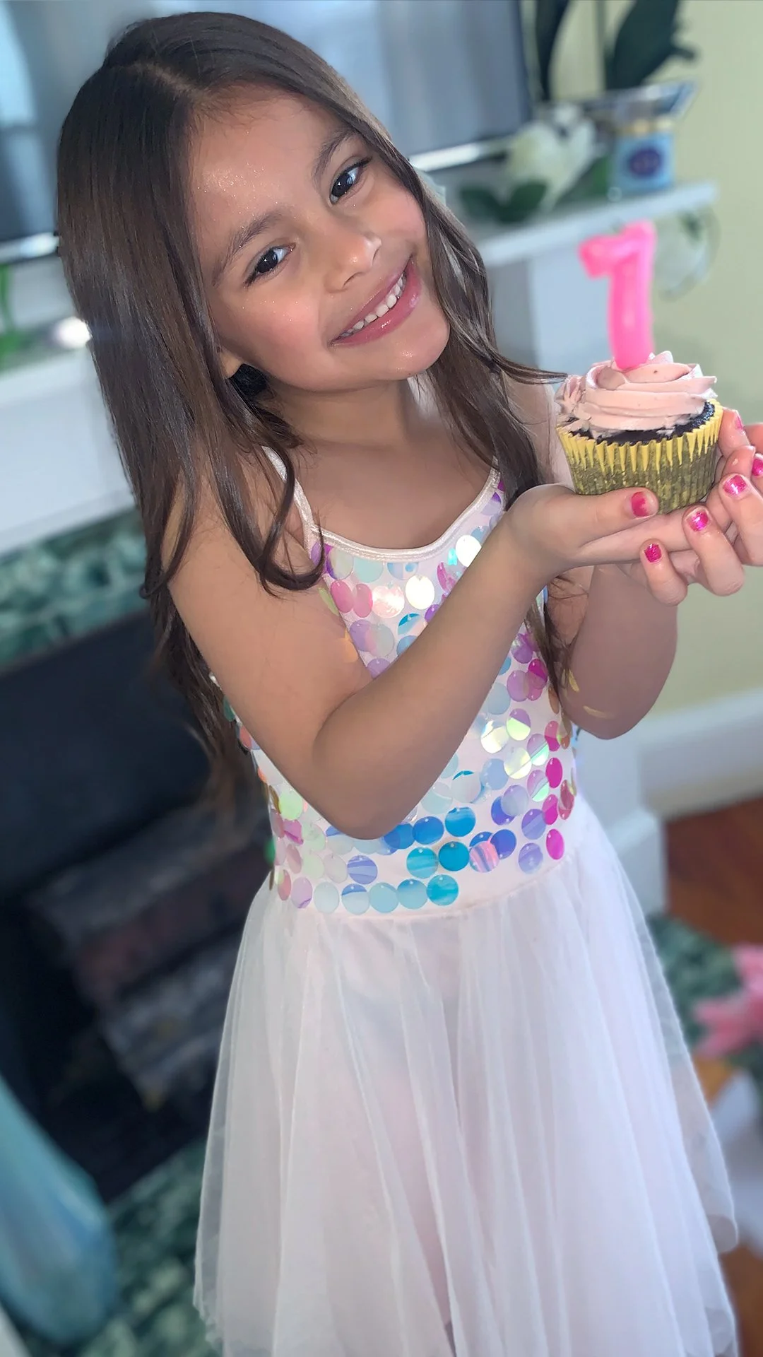 A young girl with long brown hair smiling and holding a birthday cupcake with a pink number one candle.