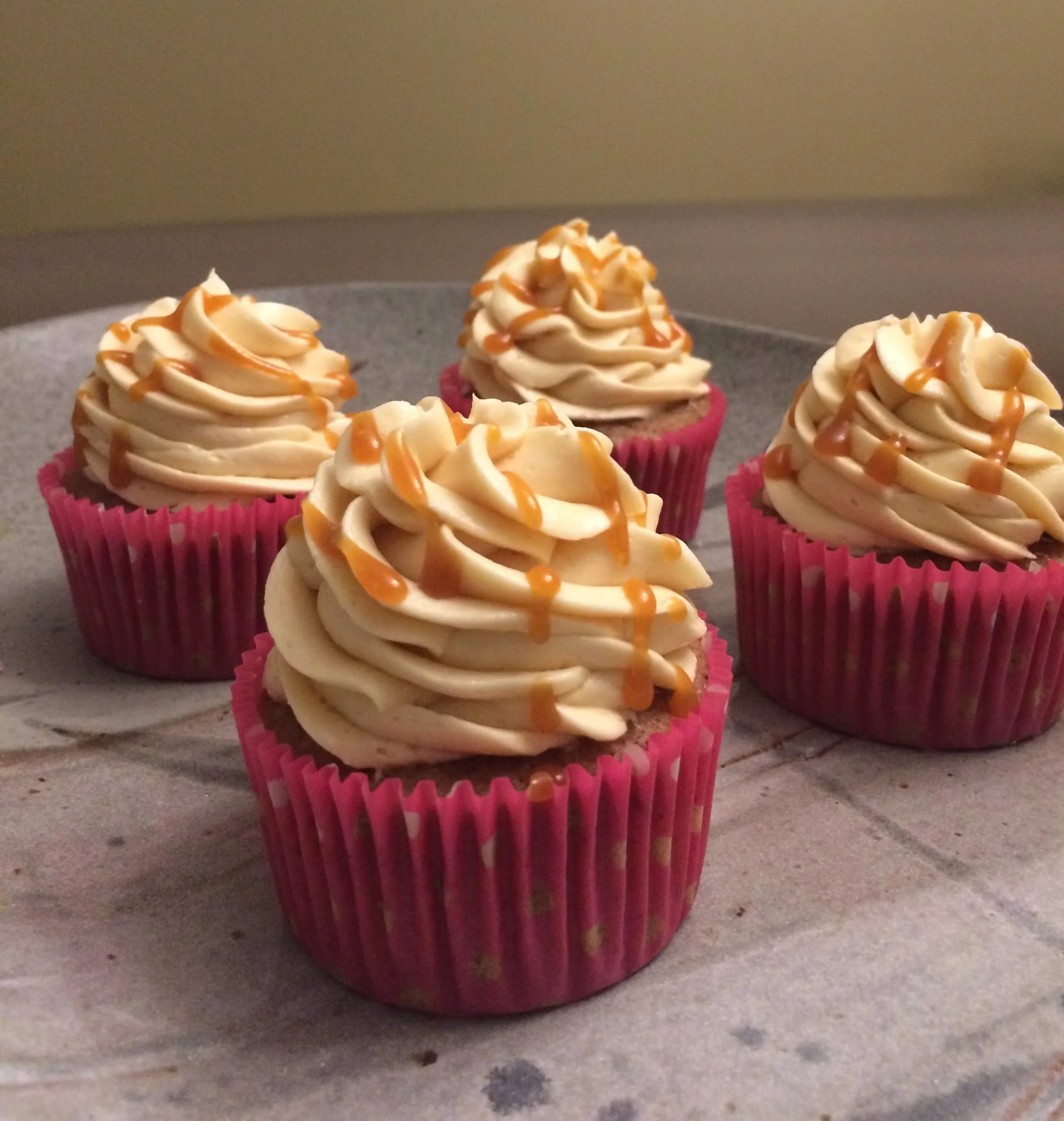 Four cupcakes with swirl frosting, drizzled with caramel, in pink cupcake liners on a gray plate.