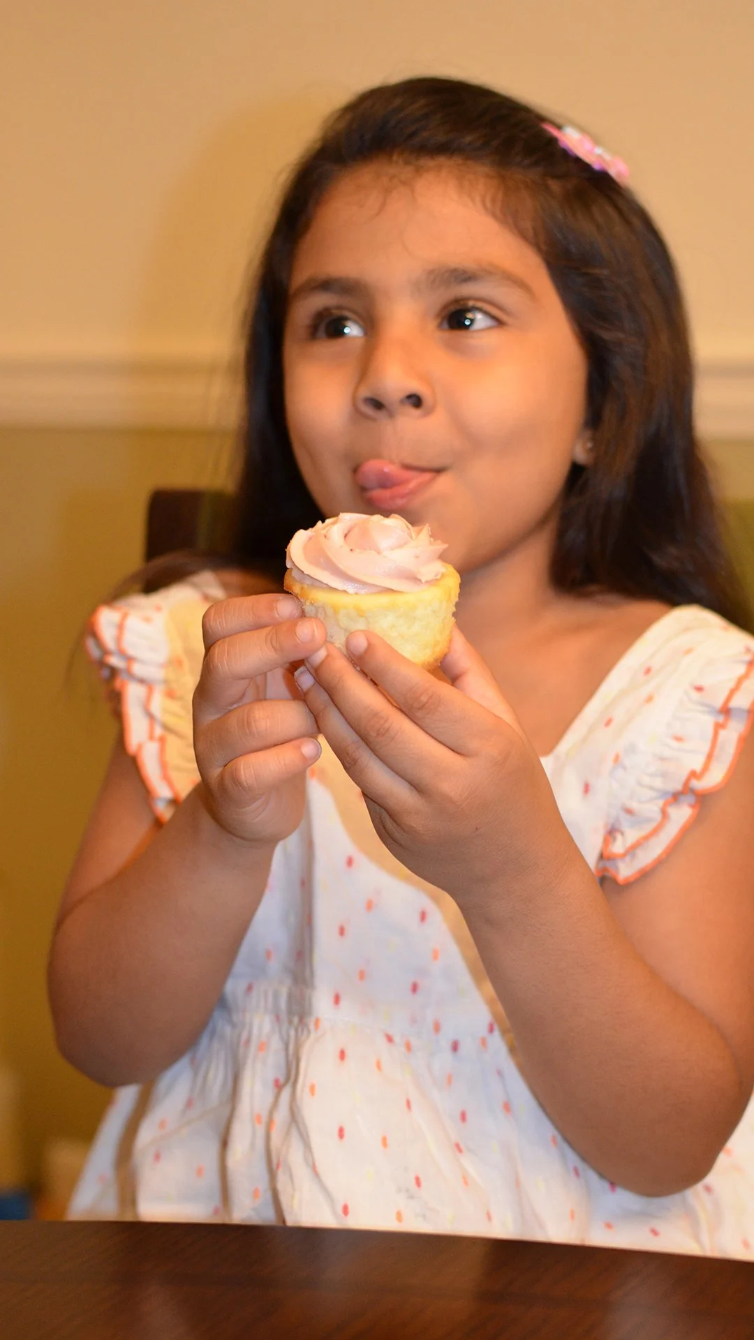 Young girl with dark hair and a pink hair clip enjoying a cupcake with pink frosting.