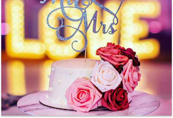 A wedding cake decorated with pink, white, and dark red roses, with a topper that reads 'Mr & Mrs,' and blurred yellow lights in the background.