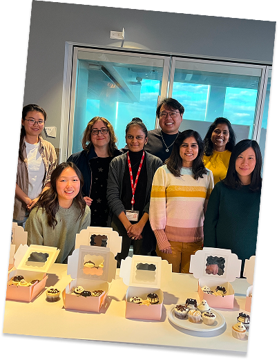A group of eight smiling women standing behind a table with decorated cupcakes in pink boxes, in a room with large glass windows showing a cloudy sky.