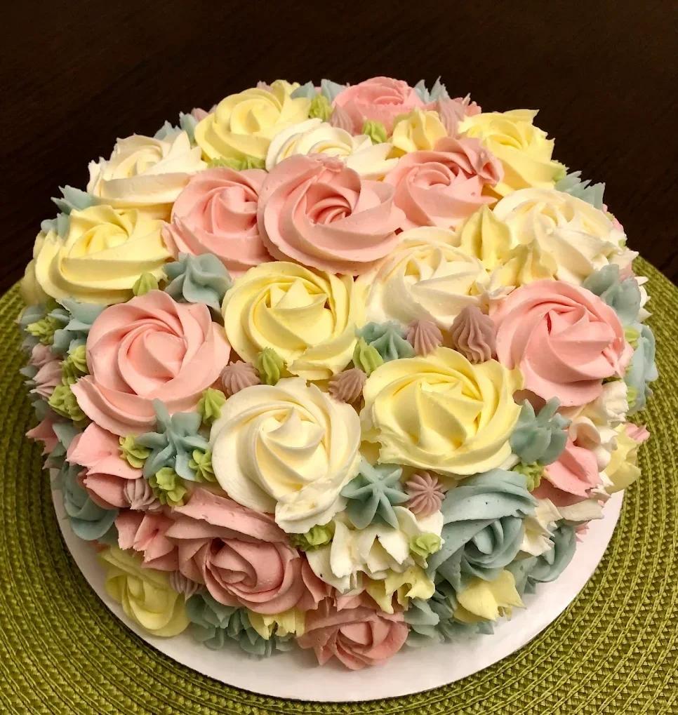 A round cake decorated with pastel-colored buttercream roses and small flower accents, placed on a green woven placemat.