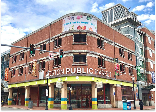 Corner view of Boston Public Market building with a large sign that says 'Bring Fresh Home Today,' traffic lights, and pedestrians on the street.