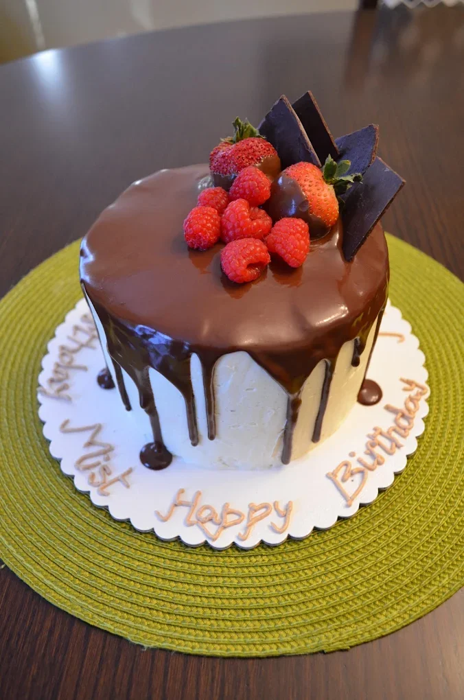 A birthday cake decorated with berries and chocolate, with chocolate ganache dripping down the sides, on a green placemat.
