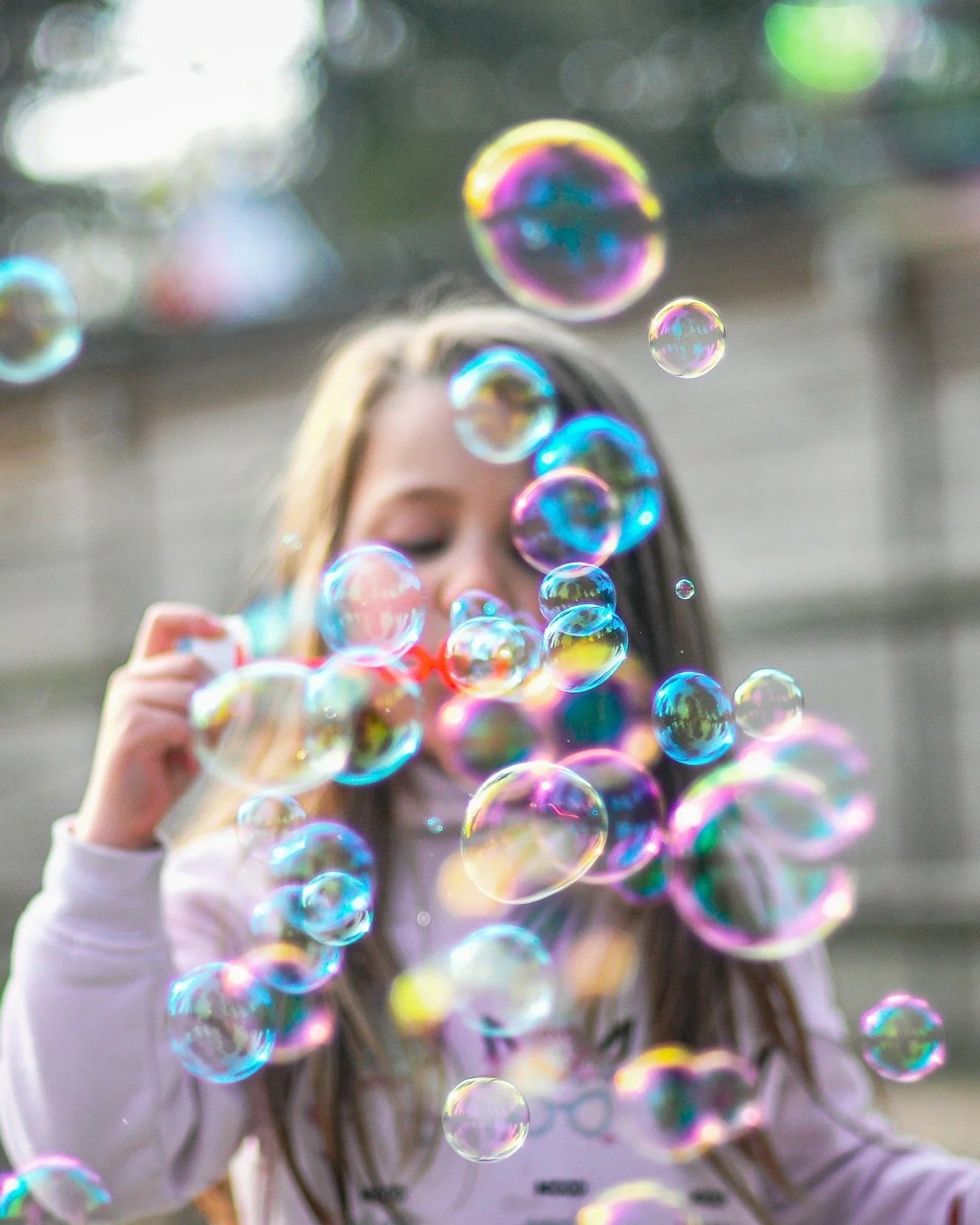 A young girl with long brown hair blowing bubbles outdoors with a blurred background.