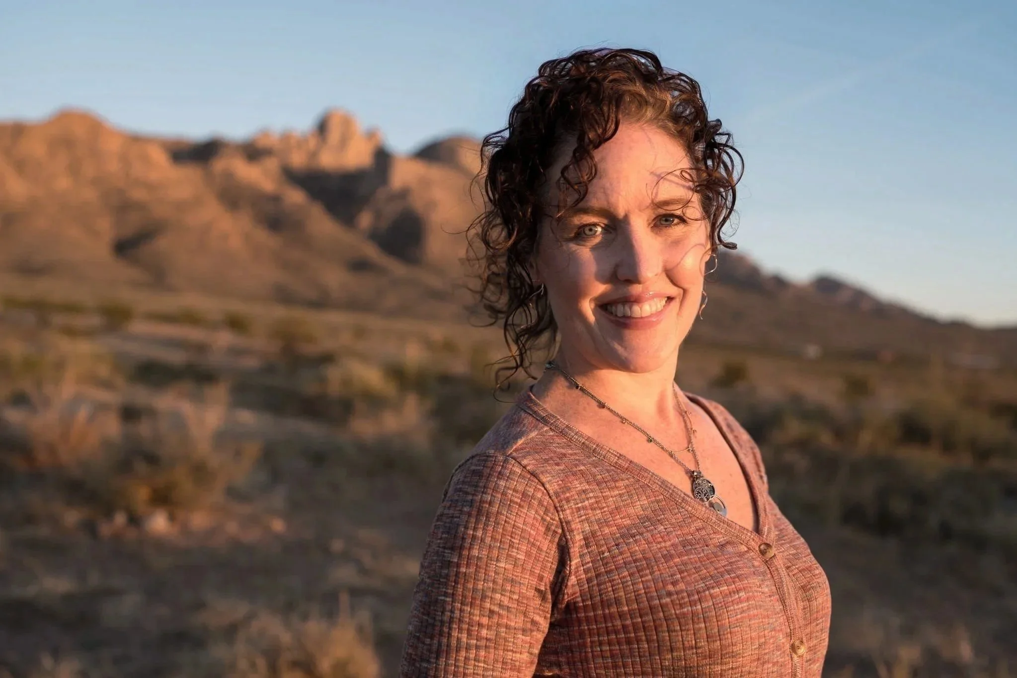 A woman with curly brown hair smiling outdoors in front of a mountainous desert landscape at sunset.