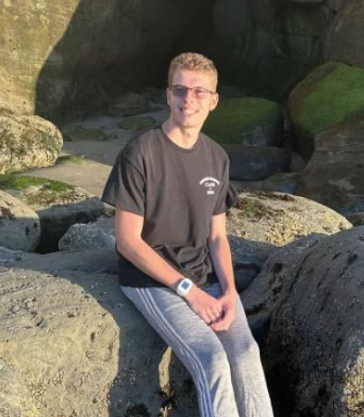 A young man with glasses, wearing a black t-shirt and gray sweatpants, sitting on a large rock at the beach with rocks and a small cave in the background.