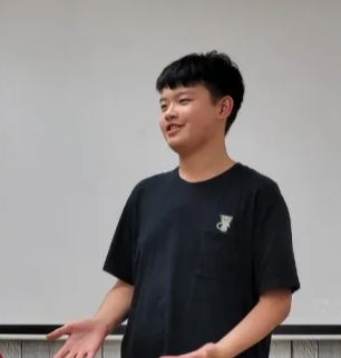 A boy standing and smiling, wearing a black T-shirt, in front of a plain light-colored background.