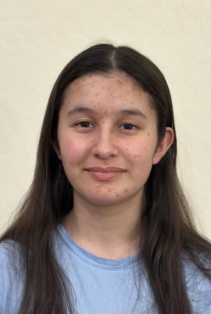 A young girl with long brown hair and light skin, wearing a light blue shirt, standing against a neutral background.
