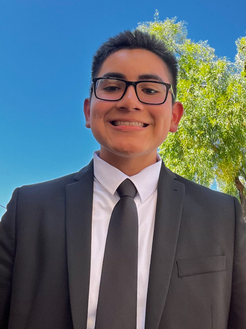 Young man smiling outdoors, wearing a black suit, white shirt, black tie, and glasses, with a large leafy tree and blue sky in the background.