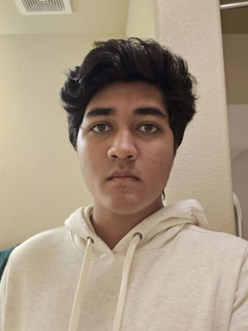 A young person with dark, wavy hair and a serious expression, wearing a cream-colored hoodie, standing indoors in front of a beige wall.