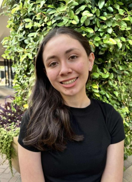 A young woman with long dark hair and a black shirt smiling outdoors in front of a leafy green hedge.