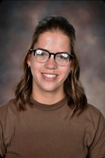 Portrait of a young woman with glasses and wavy brown hair, smiling, wearing a brown shirt.